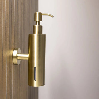 La pompe à savon Alberto Gold de yessential.fr avec support mural en acier inoxydable et fente de fenêtre verticale se fixe sur un mur en bois, sur un fond neutre.