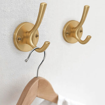 Deux élégantes patères yessential.fr Eduardo Gold Deluxe sont montées sur un mur blanc. Un crochet contient un cintre métallique avec un dessus en bois, tandis que l'autre crochet reste vide.