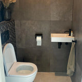 Une salle de bain moderne avec des toilettes suspendues, un petit lavabo rectangulaire avec un robinet noir et l'ensemble Armando Black de yessential.fr. Une serviette est suspendue à proximité, tandis que des accessoires élégants et des plantes séchées ajoutent de l'élégance au mur carrelé sombre.