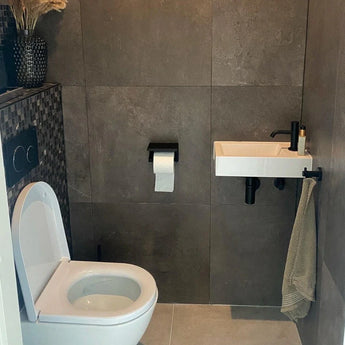 Une salle de bain moderne avec des toilettes suspendues, un petit lavabo rectangulaire avec un robinet noir et l'ensemble Armando Black de yessential.fr. Une serviette est suspendue à proximité, tandis que des accessoires élégants et des plantes séchées ajoutent de l'élégance au mur carrelé sombre.