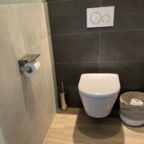 Une salle de bain moderne avec toilettes murales, boutons à double chasse et ensemble de toilettes yessential.fr Ludo Chrome (sans perceuse) avec porte-papier toilette et brosse, plus un panier tressé sur un sol en bois et des murs carrelés clair/foncé.
