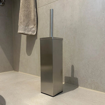 La brosse WC Velasco en acier inoxydable avec support de la marque yessential.fr, au design rectangulaire, se dresse sur un sol carrelé de couleur claire dans la salle de bain, à côté d'une serviette beige accrochée au mur.