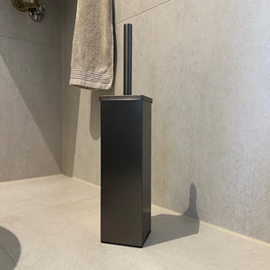Une brosse de toilette moderne et rectangulaire yessential.fr Velasco Gunmetal avec une finition brossée sur le sol carrelé de la baignoire, avec un côté beige, avec une serviette beige à proximité.