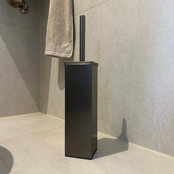 Une brosse de toilette moderne et rectangulaire yessential.fr Velasco Gunmetal avec une finition brossée sur le sol carrelé de la baignoire, avec un côté beige, avec une serviette beige à proximité.