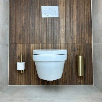 Une salle de bain moderne avec des toilettes suspendues blanches, des boiseries verticales et la garniture de toilette Cosimo Gold de yessential.fr : brosse de toilette dorée à droite et porte-papier toilette à gauche. Le bouton de chasse d'eau se trouve au dessus des toilettes.