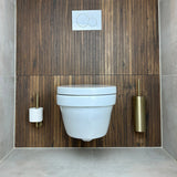 Une salle de bains moderne comprend des toilettes suspendues blanches, une paroi arrière en bois et l'ensemble de toilettes yessential.fr Lauro Gold, avec un porte-brosse de toilette doré à droite et un porte-papier toilette assorti avec rouleau à gauche.