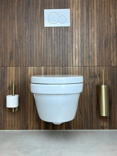 Une salle de bains moderne présente des toilettes murales blanches contre des panneaux en bois, avec à droite l'ensemble WC Fabiano Gold de yessential.fr, comprenant un porte-brosse de toilettes doré. Le sol et les murs latéraux sont carrelés en gris clair.