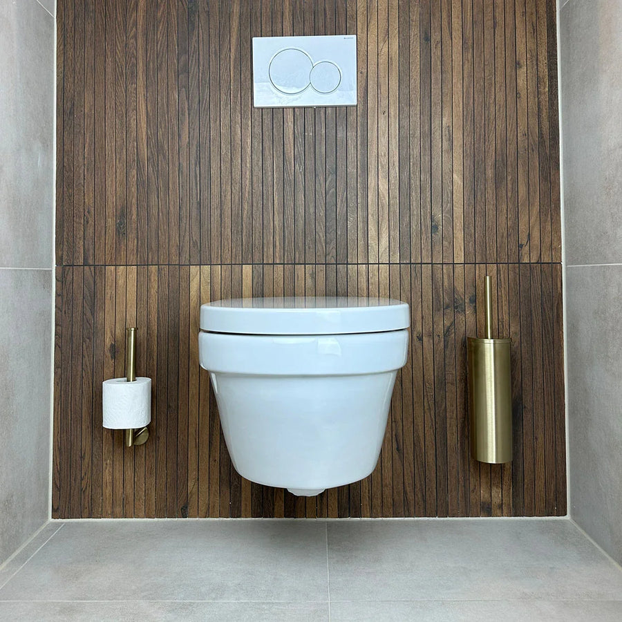 Une salle de bains moderne présente des toilettes murales blanches contre des panneaux en bois, avec à droite l'ensemble WC Fabiano Gold de yessential.fr, comprenant un porte-brosse de toilettes doré. Le sol et les murs latéraux sont carrelés en gris clair.