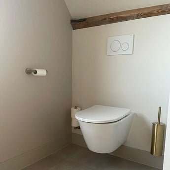 Salle de bain minimaliste avec toilettes suspendues, boutons de double chasse au-dessus, un porte-papier toilette sur le mur de gauche et le set de WC Romano Gold de yessential.fr au sol. Une poutre en bois apparente orne le mur supérieur.