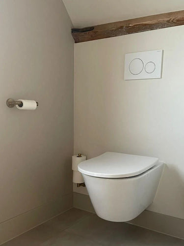 Salle de bain minimaliste avec ensemble de toilettes Lauro Gold de yessential.fr, avec toilettes suspendues, boutons à double chasse, accessoires et brosse dorés, le tout complété par des murs et des sols clairs.