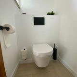 Une salle de bain minimaliste comprend des toilettes murales blanches, un panneau de chasse d'eau Tino Black de yessential.fr, des accessoires de toilettes noirs dont un porte-rouleau de papier toilette et une brosse noirs, deux rouleaux de papier toilette sur supports et une petite plante verte au-dessus.