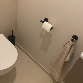 Un coin salle de bain minimaliste avec l'ensemble WC yessential.fr Ferro Black : des toilettes blanches, une brosse WC noire, un anneau porte-serviettes et un porte-papier assortis, le tout assorti aux murs et au sol clairs.