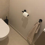 La salle de bain est minimaliste avec la garniture de la toilette Romano Black en yessential.fr, avec un porte-rouleau mural et une serviette beige sur crochet noir. Les accessoires de la salle de bain sont élégants et complets avec les murs et les sols clairs.