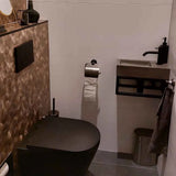 Une salle de bain moderne avec toilettes noires, carrelage mural hexagonal à structure marron, lavabo rectangulaire compact avec robinet noir, ensemble WC Mano Gunmetal de yessential.fr, sol gris, porte-papier toilette mural et poubelle en métal.