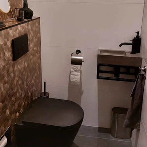 Une salle de bain moderne avec toilettes noires, carrelage mural hexagonal à structure marron, lavabo rectangulaire compact avec robinet noir, ensemble WC Mano Gunmetal de yessential.fr, sol gris, porte-papier toilette mural et poubelle en métal.