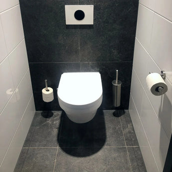 Une salle de bains moderne comprend des toilettes murales blanches, des murs et des sols carrelés foncés et l'ensemble de toilettes en acier inoxydable yessential.fr Xavo : une élégante brosse de toilettes en acier inoxydable avec support, un rouleau de papier toilette monté et un rouleau supplémentaire au sol.