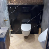 Une salle de bain moderne avec toilettes suspendues, ensemble de brosses de toilettes yessential.fr Alessino Gold, mur d'accent en marbre noir avec veines blanches, luminaires dorés, lavabo flottant à gauche et sol en bois clair à chevrons.