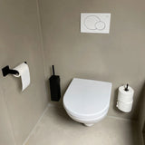 Une salle de bain moderne et minimaliste avec des toilettes murales blanches et l'ensemble Damiano Black de yessential.fr - composé d'une brosse de toilette noire et d'un porte-papier toilette avec rouleau supplémentaire - contre des murs et un sol beiges.