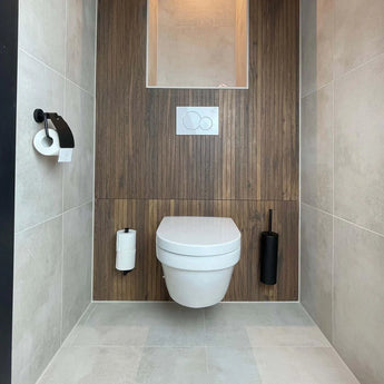Une salle de bains moderne comprend des toilettes suspendues avec des boiseries, du carrelage gris clair et l'ensemble WC Mano Black de yessential.fr - une brosse de toilette et un porte-papier noir mat - ainsi qu'une plaque de commande blanche au-dessus.