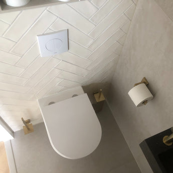 Vue de dessus d'une salle de bains moderne avec des toilettes murales blanches, des carreaux à chevrons blancs et l'ensemble de toilettes Venzo Gold de yessential.fr - comprenant un porte-brosse de toilette doré et un porte-papier toilette assorti à côté d'un lavabo noir.
