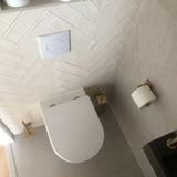Une salle de bain moderne avec les toilettes suspendues Alessino Gold de yessential.fr, une brosse WC dorée avec support assorti, un porte-papier toilette doré, un sol carrelé beige, un carrelage mural à chevrons blanc et un bouton de chasse d'eau au-dessus.