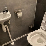 Une petite salle de bain avec un lavabo et un robinet blancs à gauche, un porte-rouleau de papier toilette en inox Tazio yessential.fr avec un rouleau et des toilettes blanches avec le couvercle ouvert à droite. Les murs et le sol sont recouverts de carrelage gris foncé.