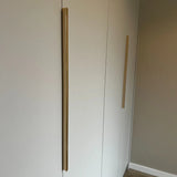 Une armoire minimaliste avec deux hautes poignées verticales Enzo Gold de yessential.fr sur des portes blanches, contre un mur neutre et un parquet en bois clair.