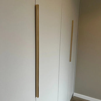 Une armoire minimaliste avec deux hautes poignées verticales Enzo Gold de yessential.fr sur des portes blanches, contre un mur neutre et un parquet en bois clair.