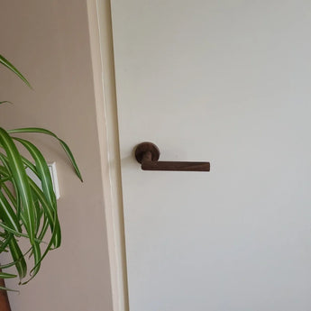 Une porte blanche avec une poignée de porte intérieure en noyer Lucio sombre et moderne de yessential.fr. À gauche, une plante à feuilles vertes et un interrupteur sont visibles sur un mur beige.