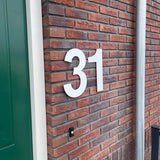 Le numéro de maison Adamo White 30cm de yessential.fr (numéro 31) est monté sur un mur de briques rouges à proximité d'une porte verte et d'un tuyau en métal gris.