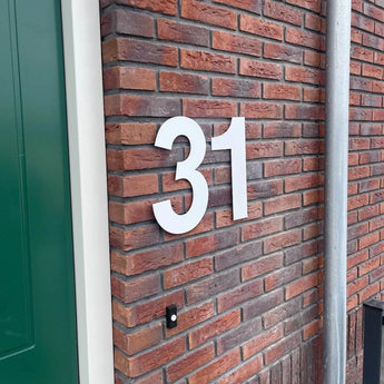Le numéro de maison Adamo White 30cm de yessential.fr (numéro 31) est monté sur un mur de briques rouges à proximité d'une porte verte et d'un tuyau en métal gris.