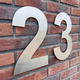 L'Adamo Stainless Steel 30cm de yessential.fr comporte un grand numéro métallique 23 avec une finition en acier inoxydable brossé, monté sur un mur de briques rouges comme un grand numéro de maison. Les chiffres sont espacés pour un look moderne et élégant.