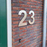 Le numéro de maison Adamo en acier inoxydable de 30 cm de yessential.fr est monté sur un mur de briques rouges à côté d'une porte verte et d'un bouton de sonnette.