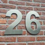 Les grands numéros Adamo de 30 cm en acier inoxydable de yessential.fr sont montés sur un mur de briques rouges et servent de numéro de maison bien visible pour une identification claire du bâtiment.