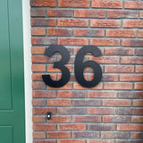 Les numéros de maison Adamo Black 30 cm de yessential.fr sont montés sous forme de numéro 36 flottant en acier inoxydable noir sur un mur de briques rouges à côté d'une porte verte.