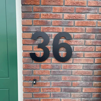 Les numéros de maison Adamo Black 30 cm de yessential.fr sont montés sous forme de numéro 36 flottant en acier inoxydable noir sur un mur de briques rouges à côté d'une porte verte.