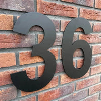 L'Adamo Black 30cm de yessential.fr est monté comme un numéro de maison flottant sur un mur de briques rouges, rendant l'adresse clairement visible.