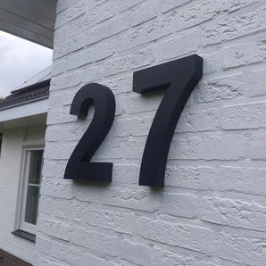 Le numéro de maison Angelo Black 30 cm de yessential.fr est monté comme un grand numéro noir frappant sur un mur de briques blanches, avec une fenêtre et un toit en arrière-plan qui soulignent le design moderne.
