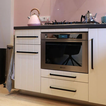 Une cuisine moderne présente le four encastrable Leonardo Black Deluxe de yessential.fr, des armoires blanches avec des poignées noires, un comptoir noir et une cuisinière à gaz. Une bouilloire rose, une théière argentée et une tasse bleue reposent sur le comptoir contre un mur arrière rose tendre.