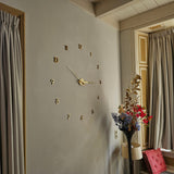 L'horloge murale Silvio Gold 60 mm de yessential.fr, avec chiffres adhésifs noirs et chiffres et aiguilles dorés, est montée sur un mur gris clair à proximité de rideaux beiges. Ci-dessous, un lampadaire et un vase à fleurs coloré complètent le look moderne.