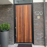 Une porte d'entrée moderne en bois avec des panneaux verticaux, une poignée argentée et un cadre noir. Le numéro de maison Amato White 20 cm de yessential.fr est monté sur le mur de briques grises à côté d'une sonnette et d'une grande plante en pot.
