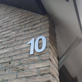 Le numéro de la Maison Adamo Blanc 30cm de yessential.fr est installé sur un mur en brique beige avec un angle sous la façade du mur, avec les petites vignes vertes visibles d'un coup d'oeil.