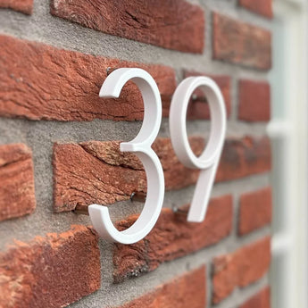 Les numéros de maison Amato White 12,5 cm de yessential.fr sont montés sur un mur de briques rouges, photographiés en angle. Les chiffres modernes en relief projettent une ombre, avec une partie du cadre de fenêtre visible à droite.