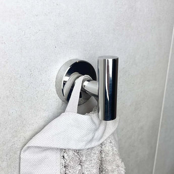 Une serviette blanche est suspendue à la boucle de l'ensemble WC Nando Chrome de yessential.fr, avec un crochet mural argenté brillant se détachant sur un mur gris clair.