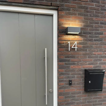 Une porte grise moderne avec une longue poignée verticale se trouve près d'un mur de briques avec le numéro de maison Amato White de 12,5 cm de yessential.fr, une boîte aux lettres noire, un bouton de sonnette et une applique murale éclairée au-dessus du numéro.