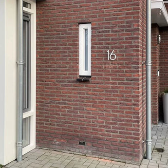 Une maison en brique rouge avec une fenêtre verticale étroite et des numéros de maison modernes. Le numéro 16, Amato Blanc 15cm est fixé au mur à côté d'un tuyau d'évacuation gris, avec des pavés au sol.
