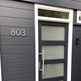 Porte d'entrée moderne gris foncé avec panneaux en verre dépoli et grande poignée, placée dans un mur assorti. Le numéro de maison 803 est illustré à gauche en Amato White 12,5 cm de yessential.fr et ajoute une touche élégante et moderne.