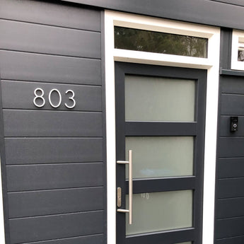 Porte d'entrée moderne gris foncé avec panneaux en verre dépoli et grande poignée, placée dans un mur assorti. Le numéro de maison 803 est illustré à gauche en Amato White 12,5 cm de yessential.fr et ajoute une touche élégante et moderne.