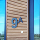 Le numéro de maison en acier inoxydable Adamo Noir 30 cm de yessential.fr agrémente une façade en bois à côté d'une fenêtre. Sous le numéro de maison noir flottant se trouve un petit interphone ou sonnette argenté. Les arbres et le ciel se reflètent dans le verre.