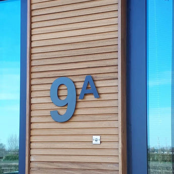 Le numéro de maison en acier inoxydable Adamo Noir 30 cm de yessential.fr agrémente une façade en bois à côté d'une fenêtre. Sous le numéro de maison noir flottant se trouve un petit interphone ou sonnette argenté. Les arbres et le ciel se reflètent dans le verre.